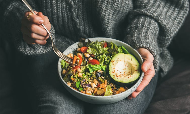 A person in a grey jumper holds a bowl of greens on their lap