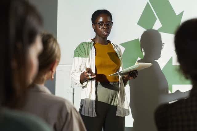 A person stands in front of a green recycling sign speaking to an audience