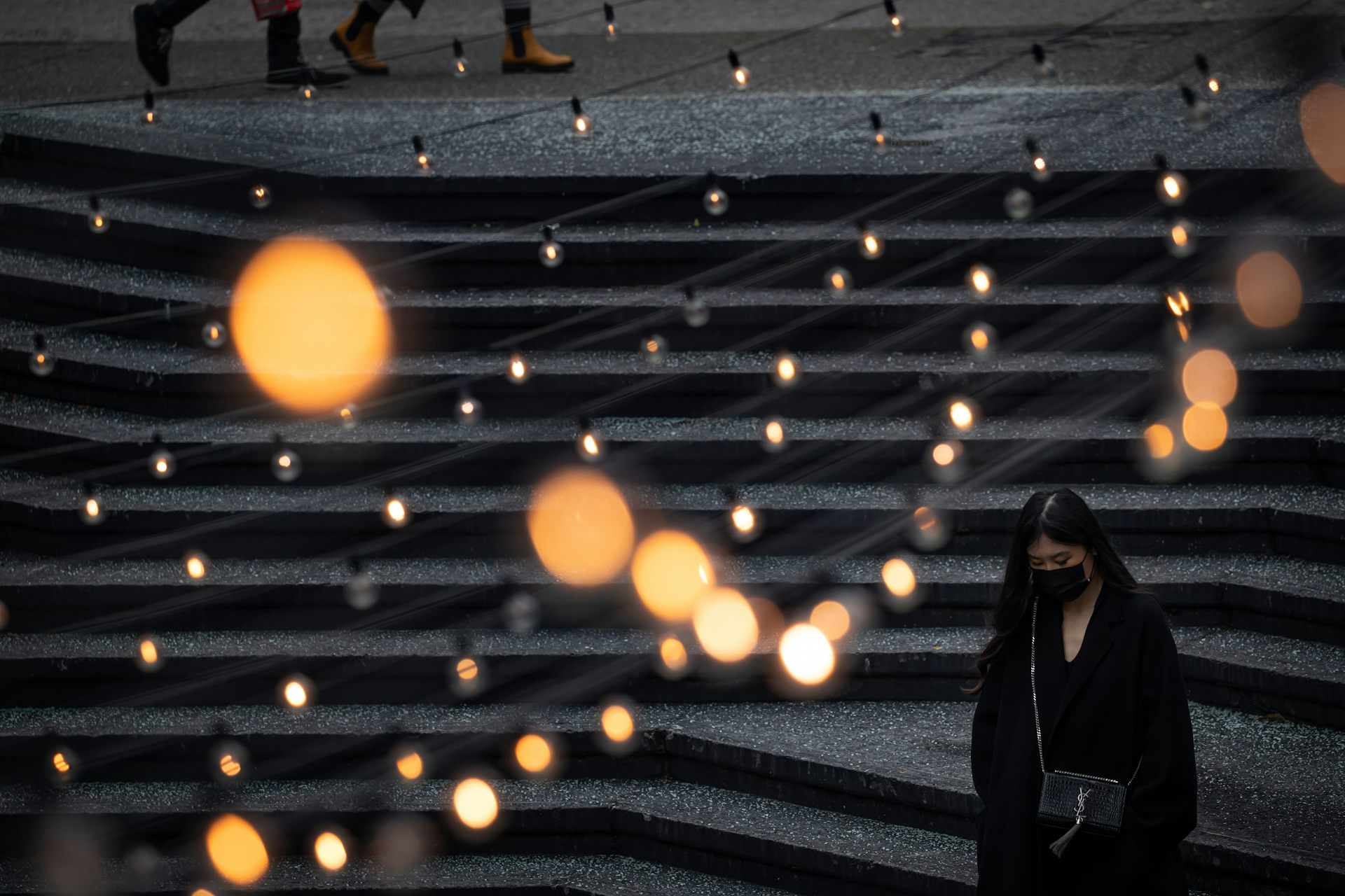 A woman wearing a mask walks through a public plaze with lights overhead.
