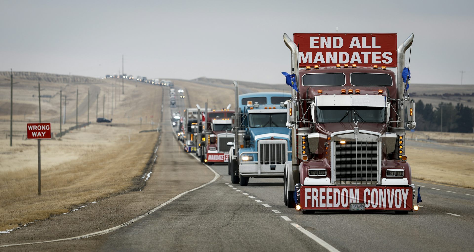A line of trucks travels down a highway, with the lead one carrying a banner that reads End All Mandates.