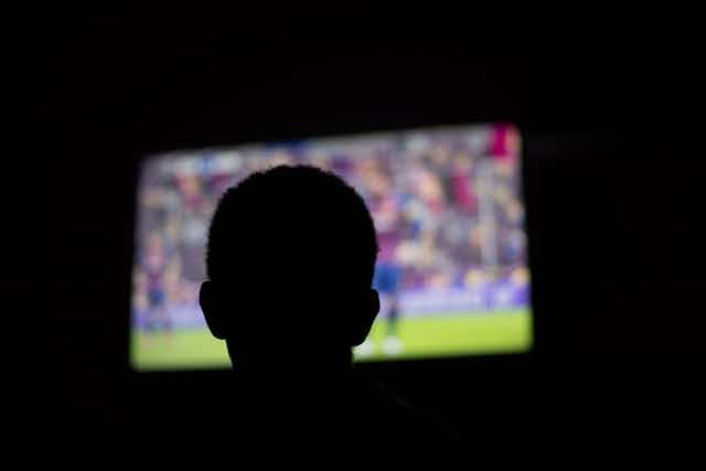 A darkened room, the silhouette of a young person watch a big screen television.