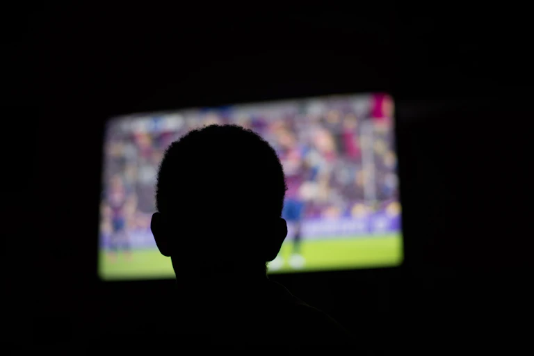 A darkened room, the silhouette of a young person watch a big screen television.