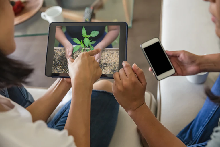 Close up shot of two female friends watching online gardening tutorial from digital tablet. Both are sitting on the sofa at living room. One woman pointing to digital tablet and another one holding her smartphone.
