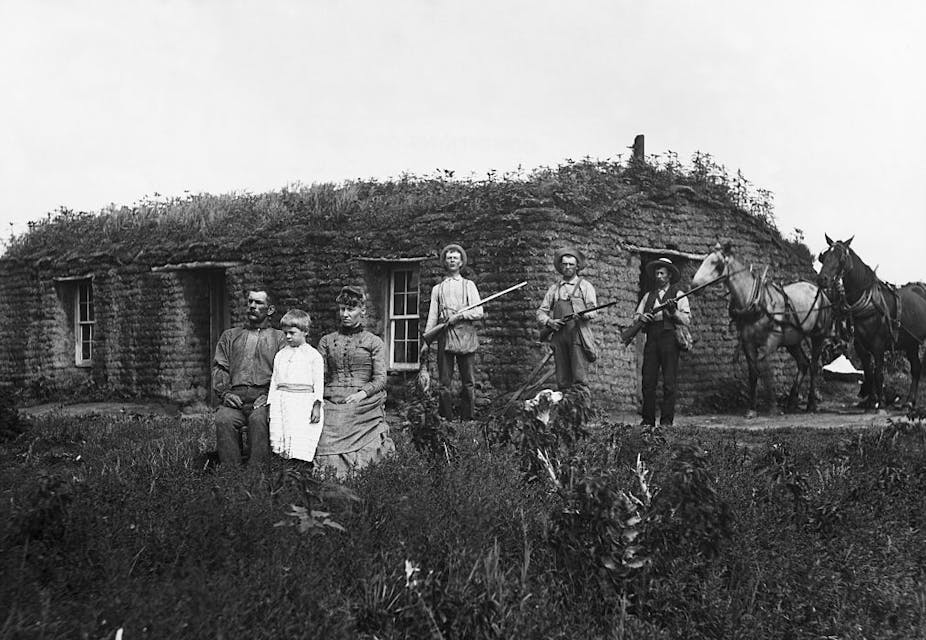 A historic black and white photograph shows four men, a woman, and a child in front of a sod house, with three of the men clutching rifles.