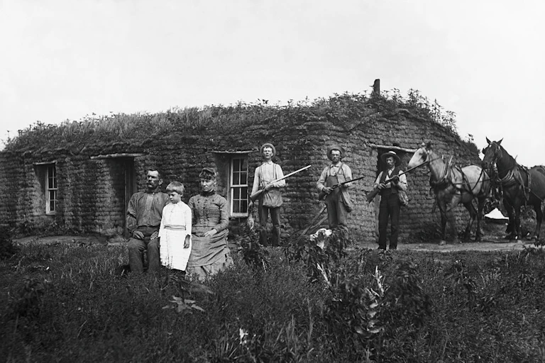 A historic black and white photograph shows four men, a woman, and a child in front of a sod house, with three of the men clutching rifles.