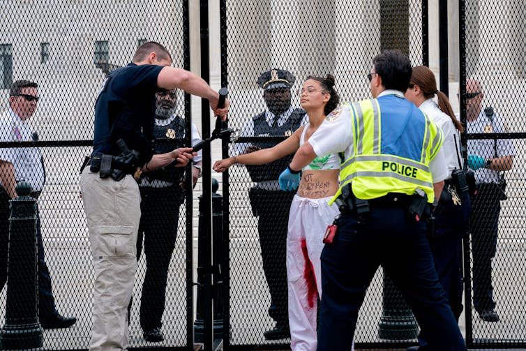 A person in white pants streaked with red on the inside is handcuffed to a fence outside the Supreme Court. Police are seen removing their handcuffs.