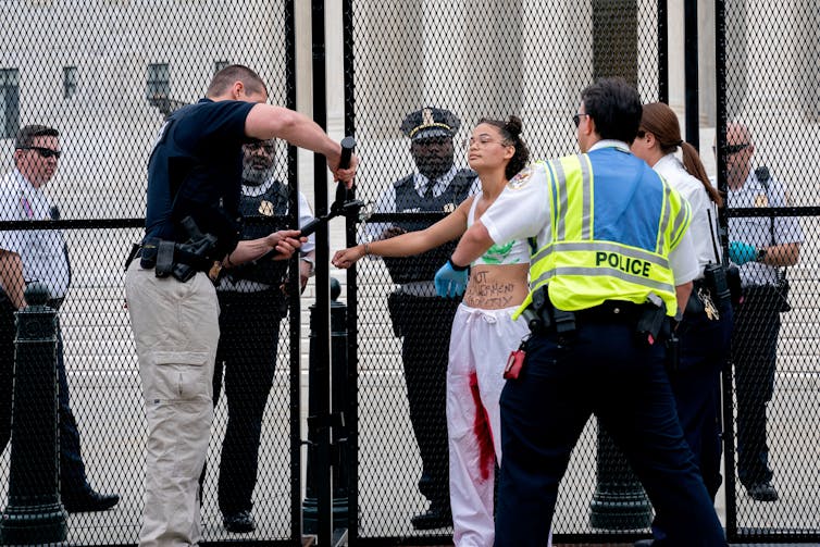 Una persona con pantalones blancos con rayas rojas en el interior está esposada a una valla frente a la Corte Suprema.  Se ve a la policía quitándose las esposas.