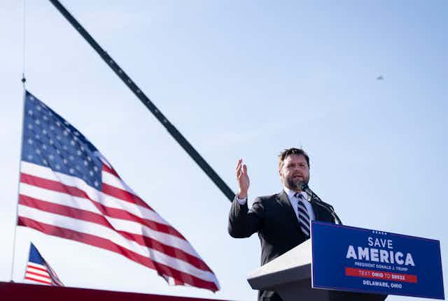 Man speaks at lecturn flanked by American flags.