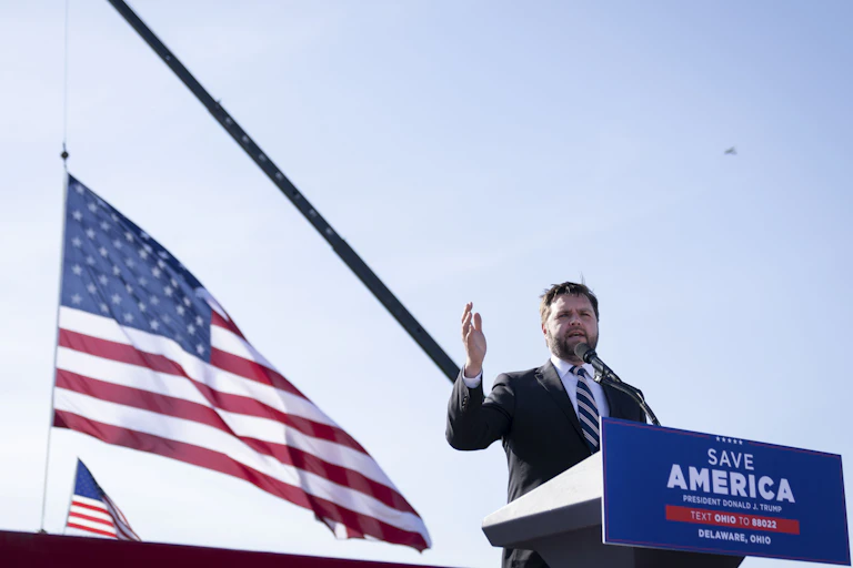 Man speaks at lecturn flanked by American flags.