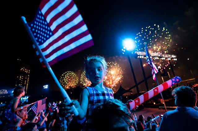 A white girl with blond hair holds a US flag as big fireworks explode in the night sky behind her