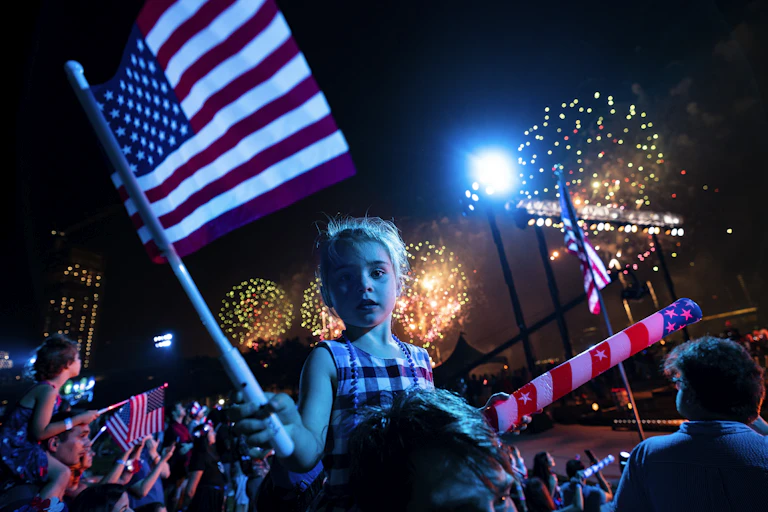 A white girl with blond hair holds a US flag as big fireworks explode in the night sky behind her