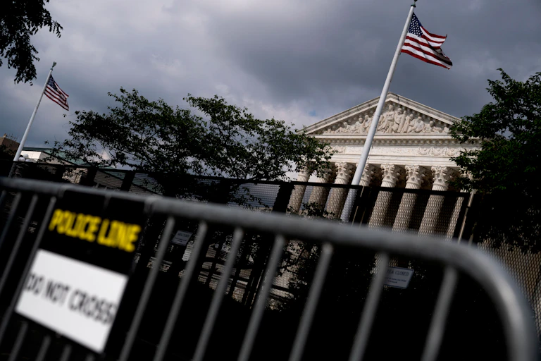 Supreme Court is shown behind fencing, with 2 American flags on a cloudy day.