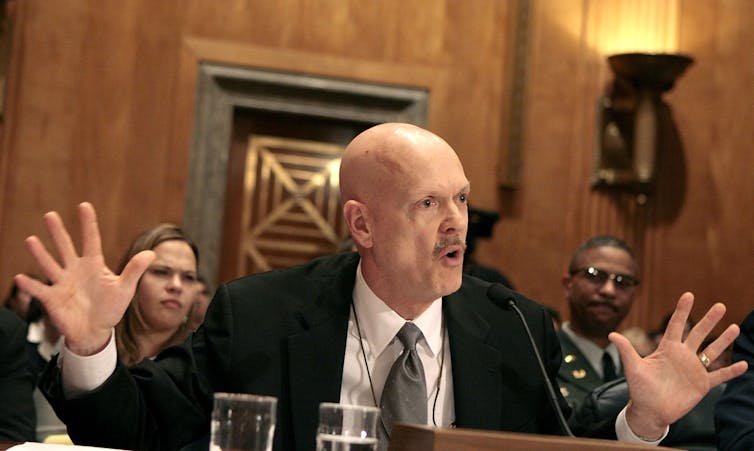 A man in a dark suit, white shirt, gesturing with both his hands out and talking to someone across the room.