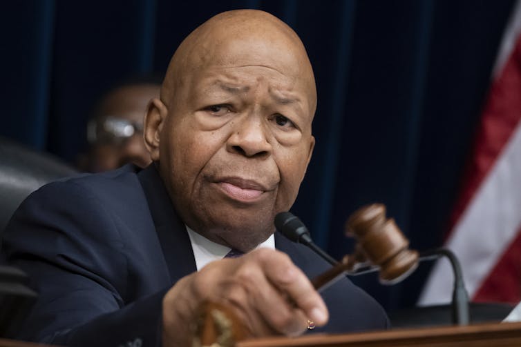 A serious-looking man in a blue suit bangs a gavel on a desktop.