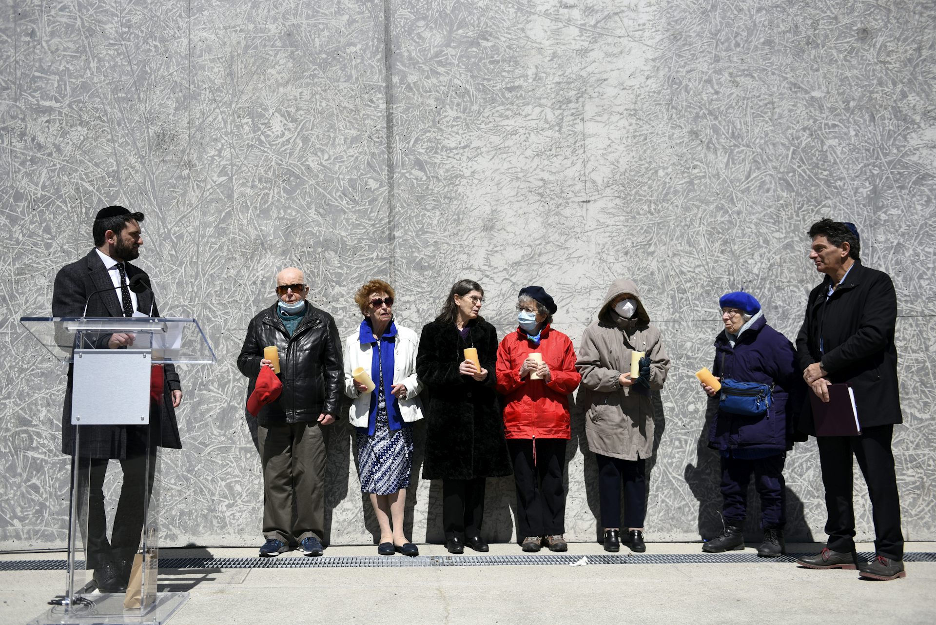 A group of older people stand leaned up against a wall holding candles