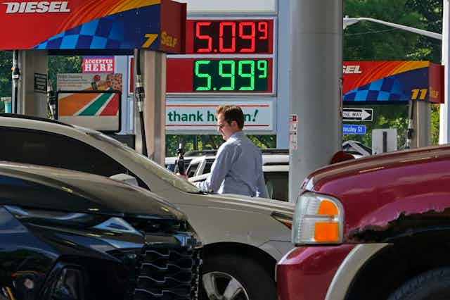 A white man with sunglasses holds a gas pump next to cars at a gas station advertising price of $5.09 a gallon