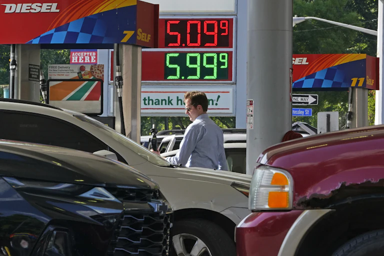 A white man with sunglasses holds a gas pump next to cars at a gas station advertising price of $5.09 a gallon