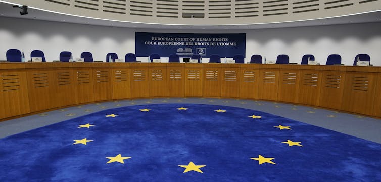Interior Of The Ecthr With An Eu Flag Carpet Surrounded By A Curved Table And A Row Of Empty Seats.
