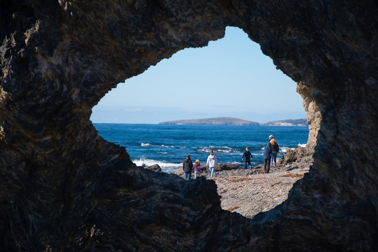 people walking along coastline viewed through hole in rock