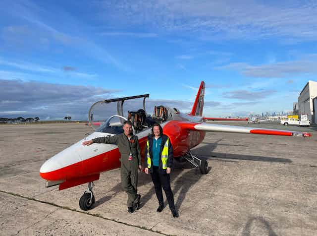 Photo of a man and a woman standing in front of a small red and white aeroplane on a runway.