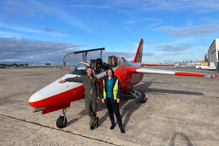 Photo of a man and a woman standing in front of a small red and white aeroplane on a runway.