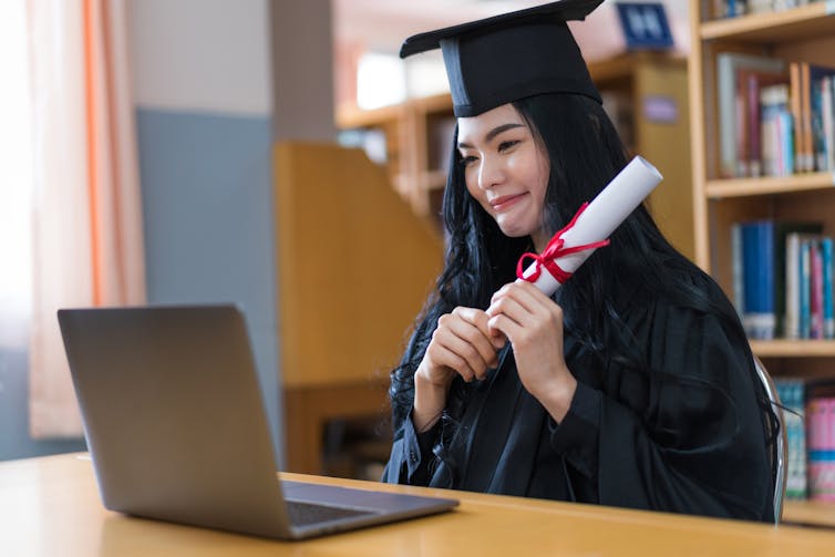 A young graduating woman sits in front of her laptop in her robe, hat and certificate in hand