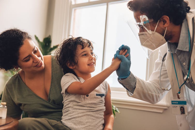 A doctor gives a child, sitting on his mother's lap, a high five after receiving a vaccination.