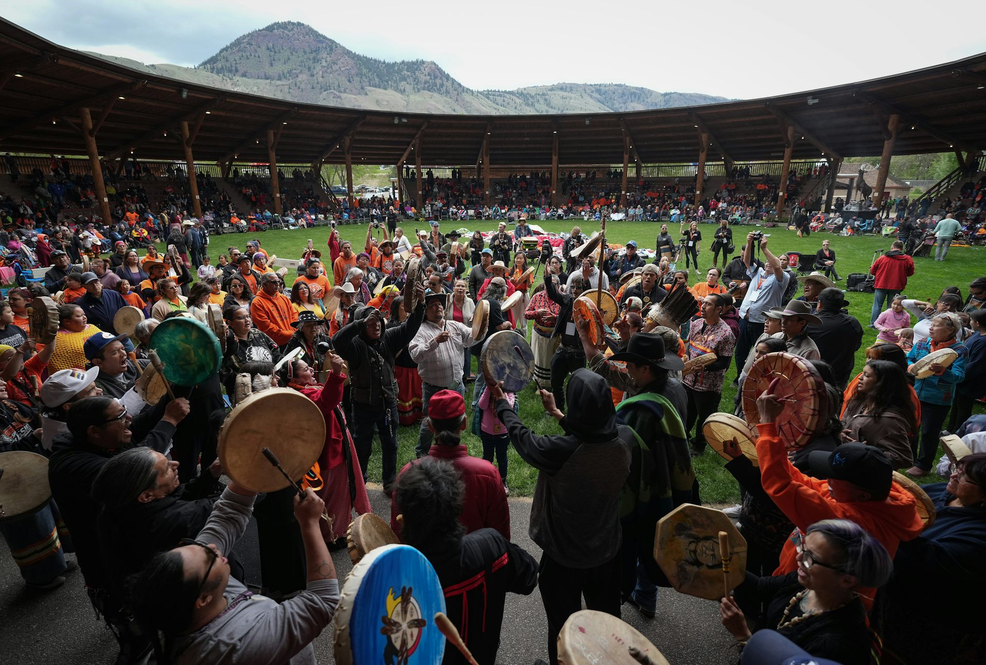 Drummers beat their drums at a large gathering with mountains in the background.