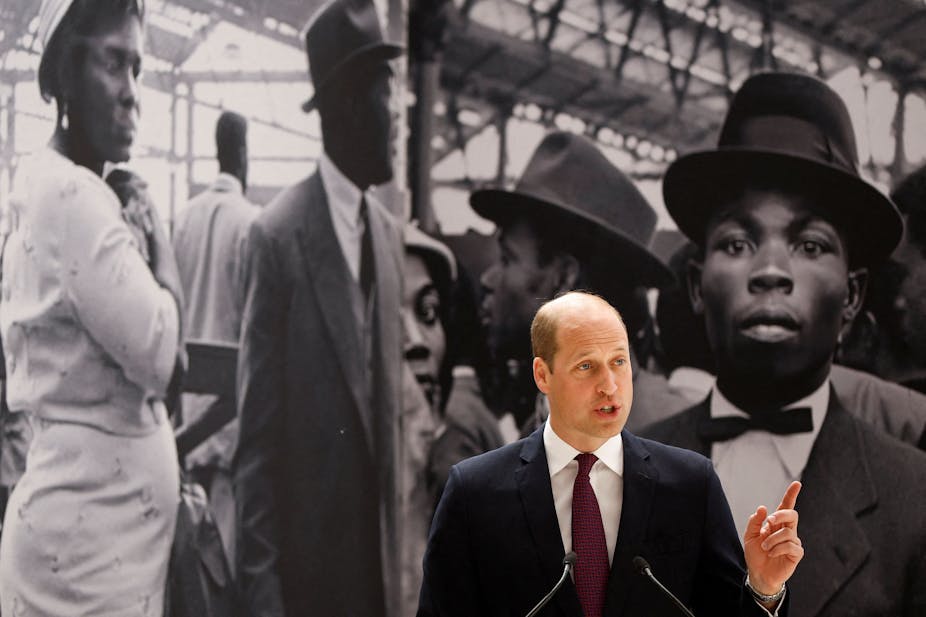 A white man is speaking in front of a large photograph of a well-dressed black woman  and two black men wearing hats and business suits. 