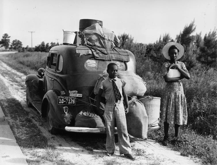 A black boy dressed in a shirt and tie stands near a black woman in front of car with their belongings tied at the top.