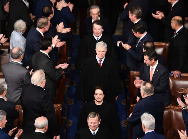 Supreme Court justices, including Elena Kagan and Brett Kavanaugh, walk in a single file line through a crowd of applauding people wearing suits and formal clothing.