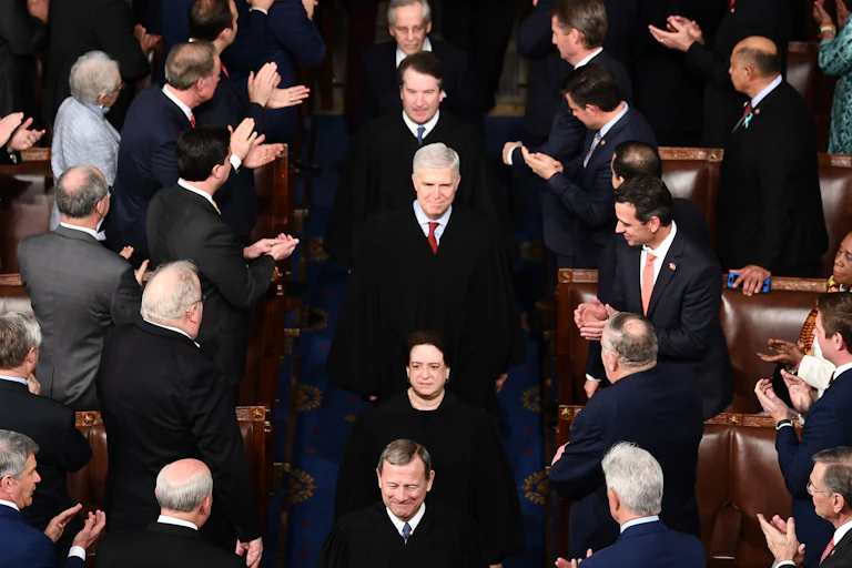 Supreme Court justices, including Elena Kagan and Brett Kavanaugh, walk in a single file line through a crowd of applauding people wearing suits and formal clothing.