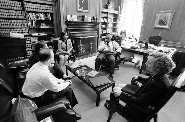 A black and white photo shows Clarence Thomas seated in an office, surrounded by young people in formal clothing.