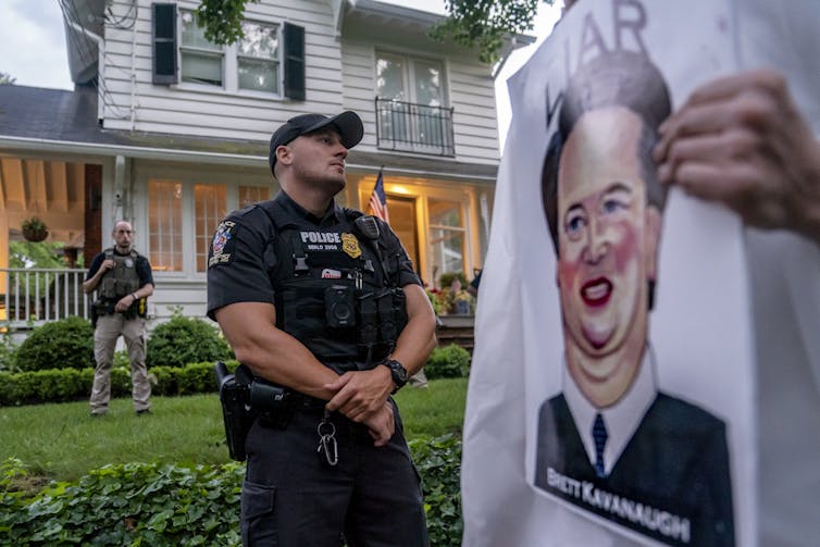 A police officer is seen standing outside a suburban home, next to a large poster that shows a drawing of Brett Kavanaugh's face, with the word liar above it