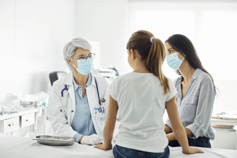 Masked female pediatrician talking with young girl and her mother in an examination room at a hospital.