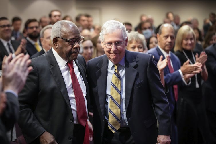 An elderly Black and an elderly white man stand next to each other as onlookers applaud.