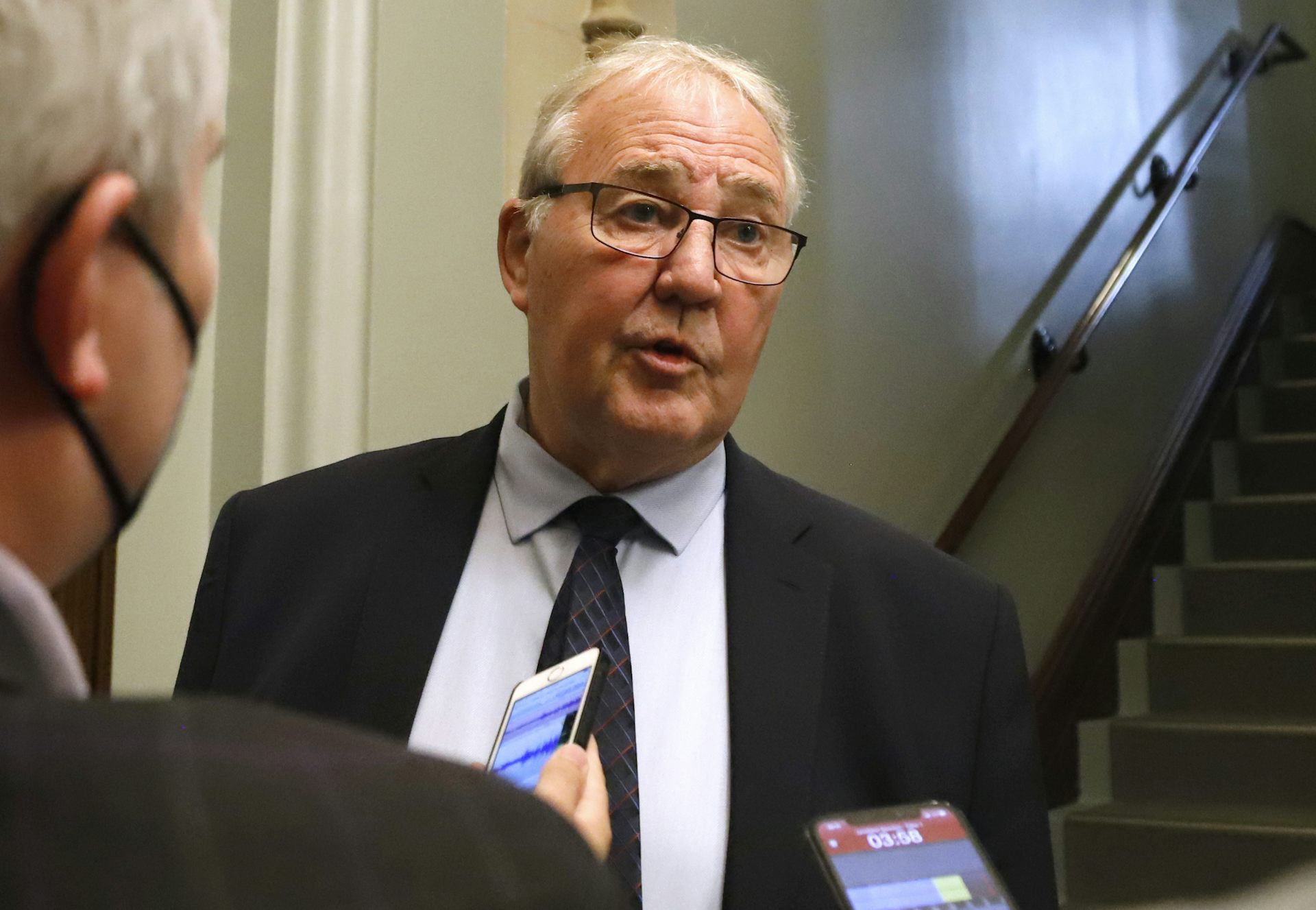 An old man wearing glasses stands as two phones are pointed at him