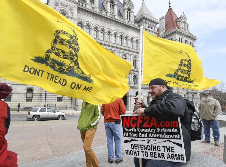 A man with a sign advocating gun rights, near two yellow 'Don't tread on me' flags and in front of a large building.