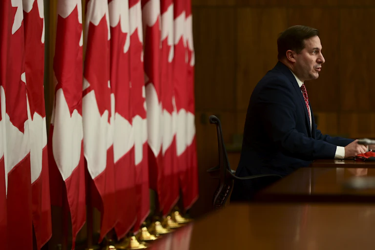 A man sitting behind a desk with a row of Canadian flags standing behind him