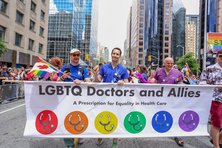 People wearing medical scrubs march in a parade carrying a banner reading LGBTQ Doctors and Allies, with rainbow coloured circles with stethoscopes in them