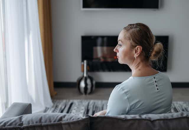 A woman sitting on a sofa seen in profile from behind, looking toward a window looking sad