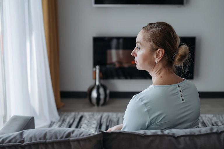 A woman sitting on a sofa seen in profile from behind, looking toward a window looking sad
