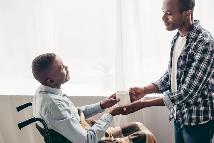 A young man standing and handing a mug to an older man seated in a wheelchair