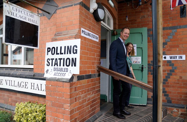 A man and woman walk out of a village hall, a brick building with Polling Station signs on the side