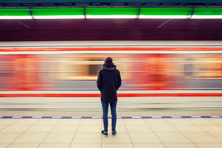 Seseorang berdiri di peron stasiun kereta api, hanya punggungnya yang terlihat, menghadap kereta api yang melintas