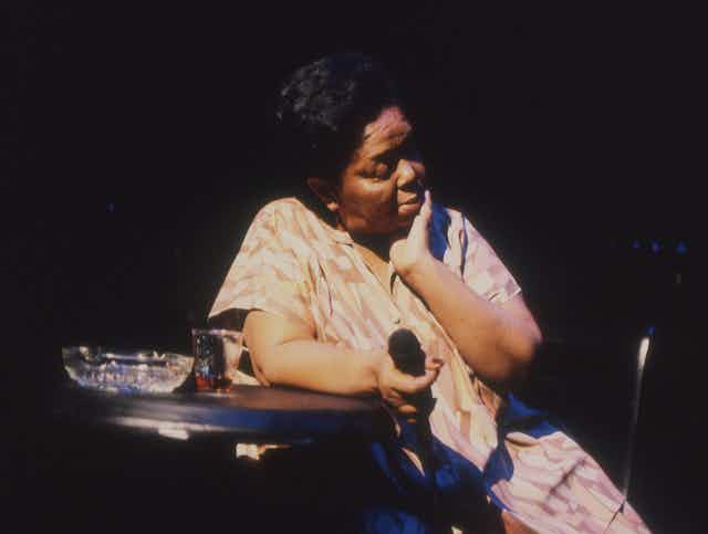 A woman at a table, a microphone in her hand, a drink and ashtray on the table, caught in stage light.