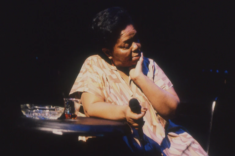 A woman at a table, a microphone in her hand, a drink and ashtray on the table, caught in stage light.