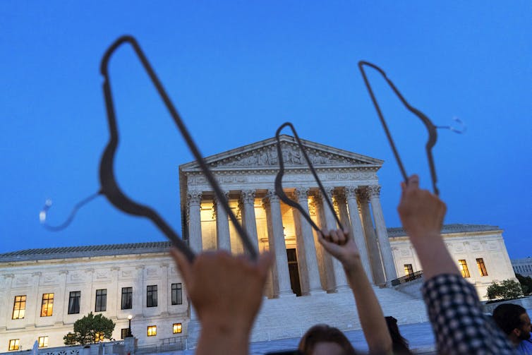 Demonstrators hold coat hangers in the air with the Supreme Court building in the background at dusk.