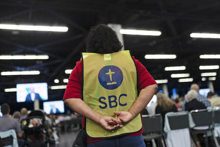 A man with a jacket that says SBC on its back stands during a meeting.