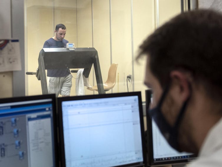 A young man in shorts walks on a treadmill with a towel beside him in a glass-enclosed room while a scientist monitors his body temperature and other conditions on computer screens on the other side of the glass.